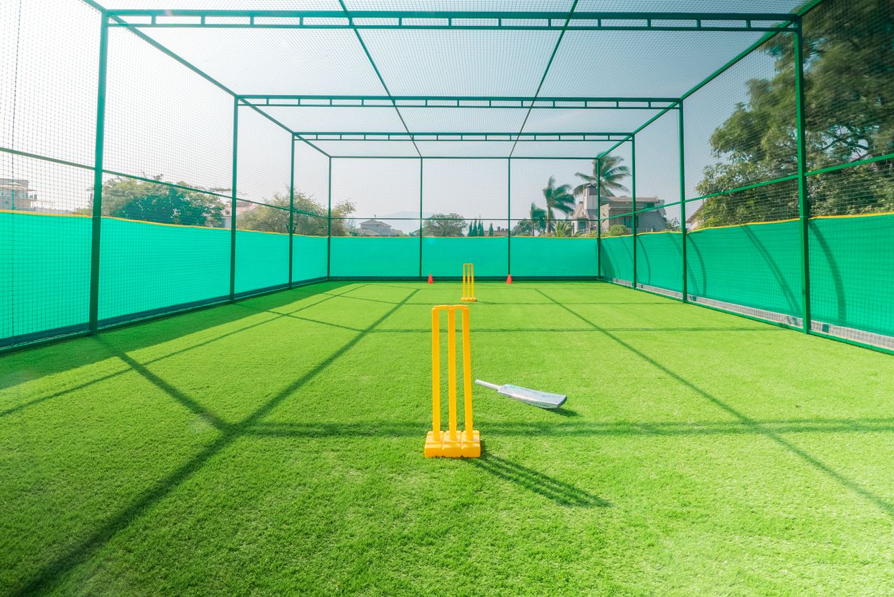 gallery-2 A cricket practice net with bright yellow stumps on a sunny day, ideal for training.