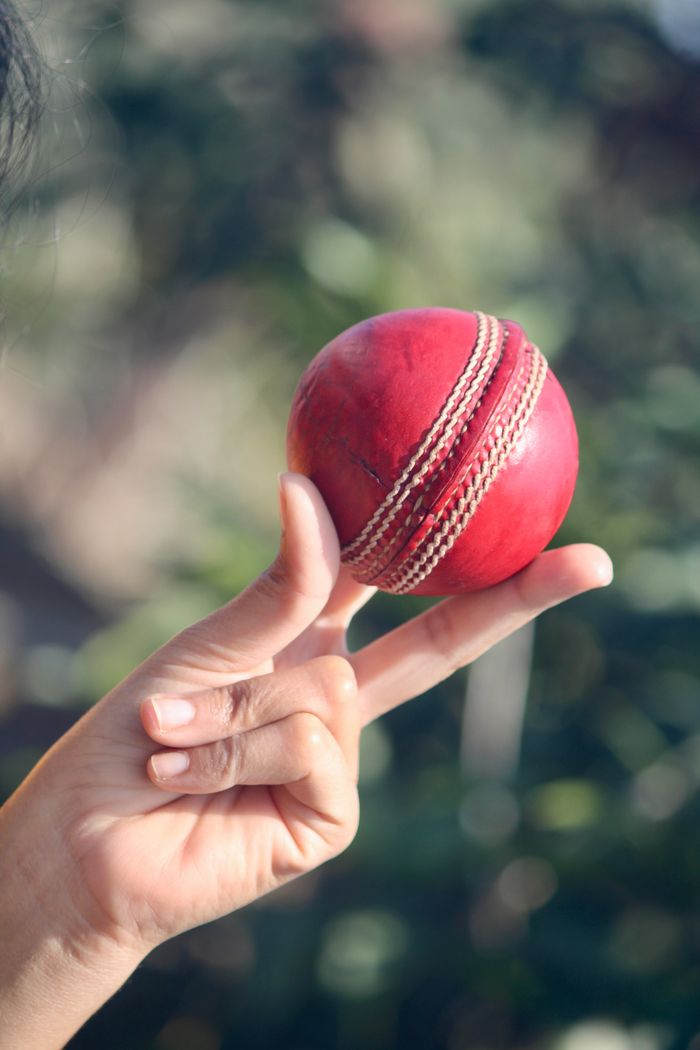 Detailed close-up of a hand skillfully holding a red leather cricket ball outdoors.