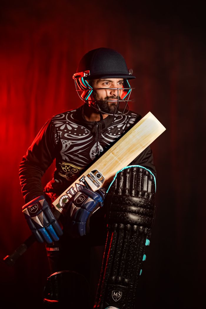 Cricket player in full gear holding a bat against a red backdrop, ready for action.